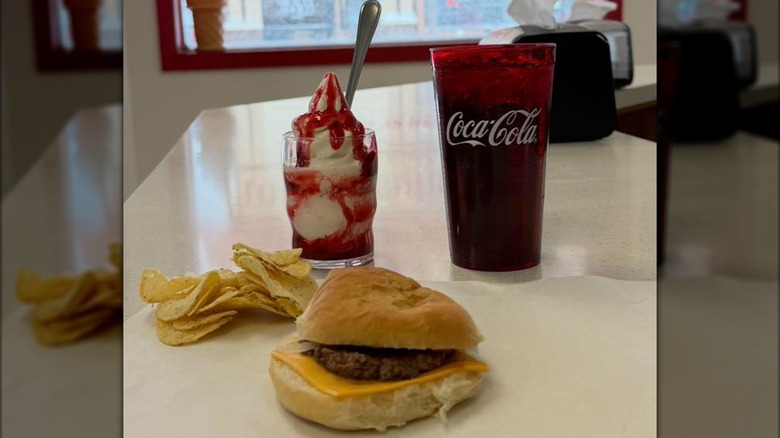 Burger with cheese on paper with chips, plastic glass of Coke, and a strawberry sundae