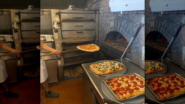 Person holding a pizza by a brick oven with two more cooked pizzas on trays nearby