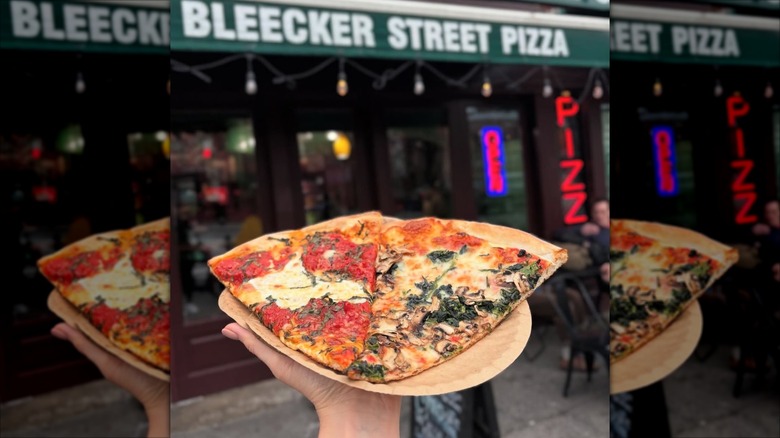 Hand holding two slices of pizza on a paper plate in front of the Bleecker Street Pizza storefront