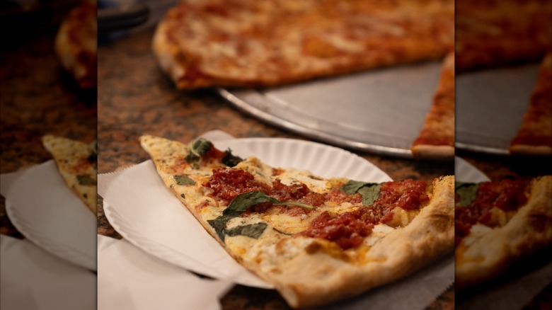 Slice of pizza on a paper plate with a whole pie (missing a slice) blurred in the background