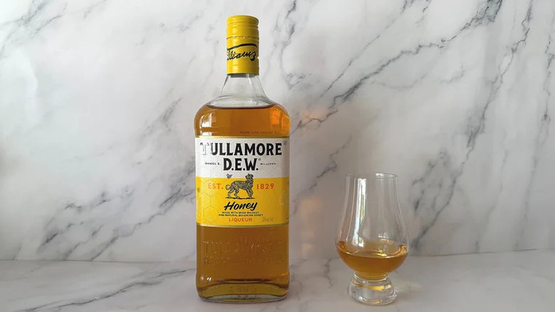 A bottle of Tullamore D.E.W Honey next to a whiskey glass partially filled with the liqueur, against a white marble background