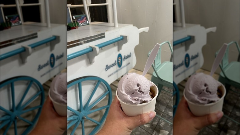 Hand holding scoop of purple ice cream in bowl with ice cream cart in background