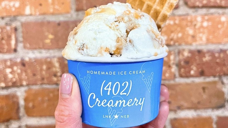 Hand holding cup of vanilla ice cream with mix-ins in front of brick wall