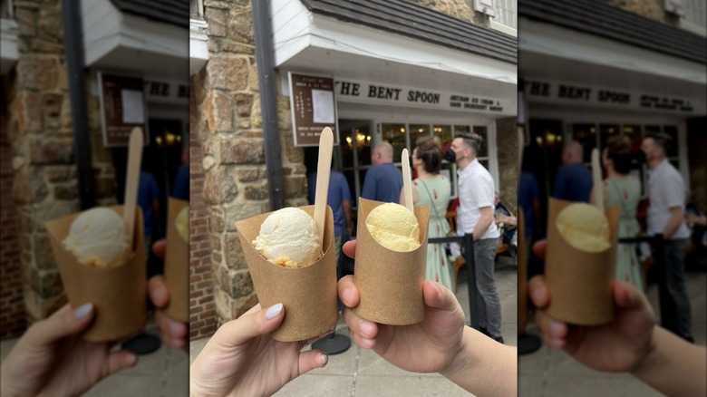 Two hands holding paper cups of ice cream in front of The Bent Spoon storefront