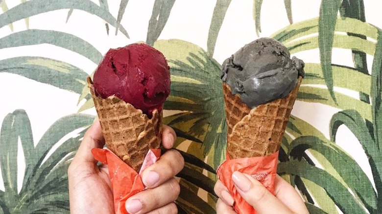 Two hands holding cones of ice cream with different flavors in front of mural