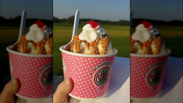 Hand holding cup filled with ice cream, topped with peanut butter sauce, whipped cream, and cherry