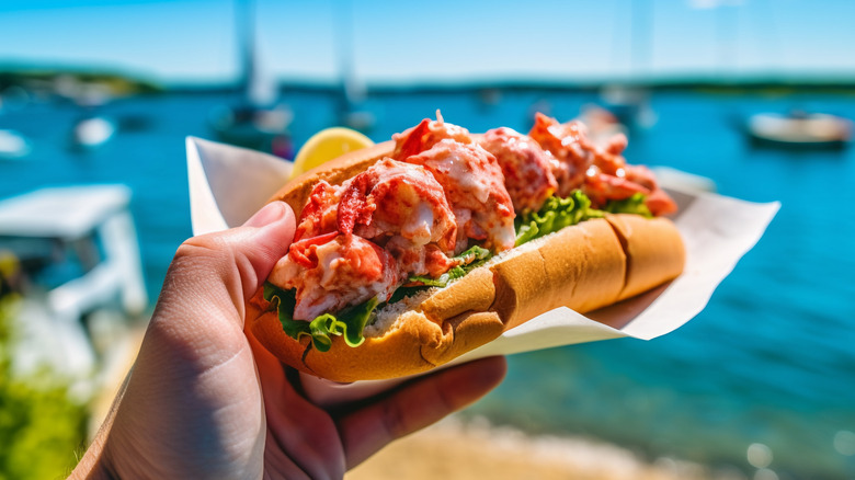Hand holding a lobster roll with the ocean in the background