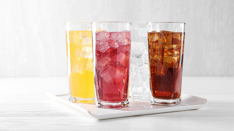 Four glasses of soda with ice cubes in tall glasses set against a white background