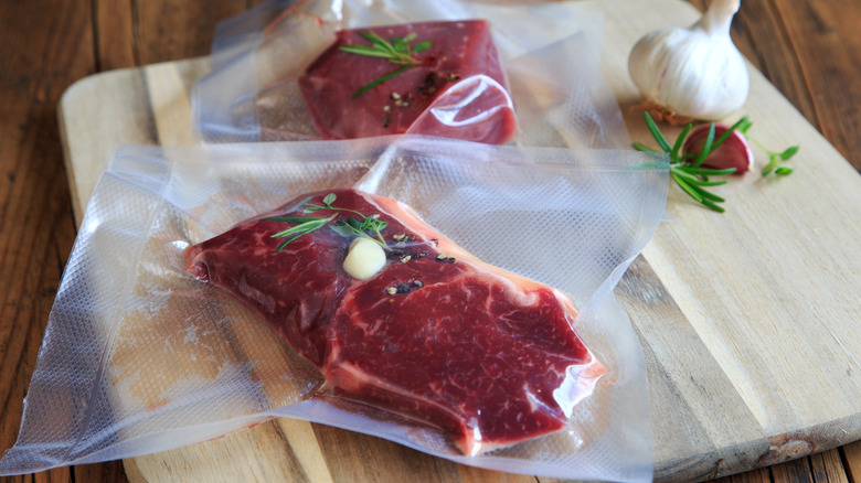 Two steaks vacuum sealed in plastic with garlic and herbs sitting on wooden board