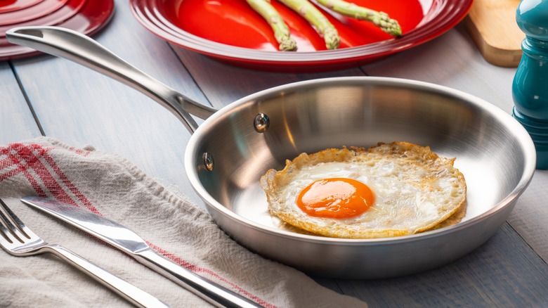 A fried egg in a stainless steel pan beside a fork and knife on a dish towel.