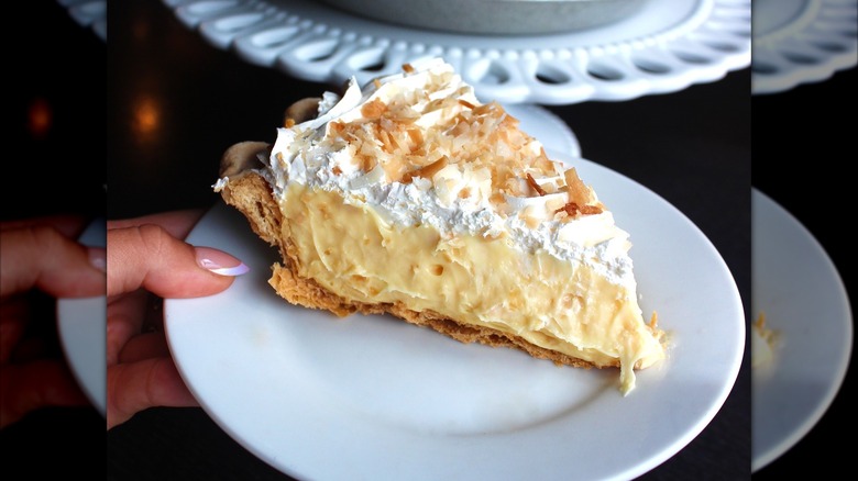 Hand holding white plate with slice of coconut cream pie