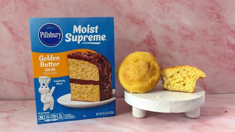 A box of Pillsbury Moist Supreme Golden Butter cake mix in front of a pink marbled background with a cupcake displayed next to it
