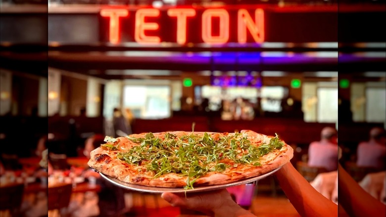 Hands holding a wood-fired pizza with greens on a tray in restaurant in front of neon Teton sign