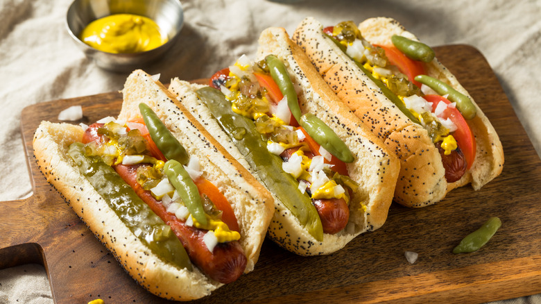 Three Chicago-style hot dogs on a wooden board, garnished with sports peppers, pickle spears, onion, and mustard, with a bowl of yellow mustard sitting in the background
