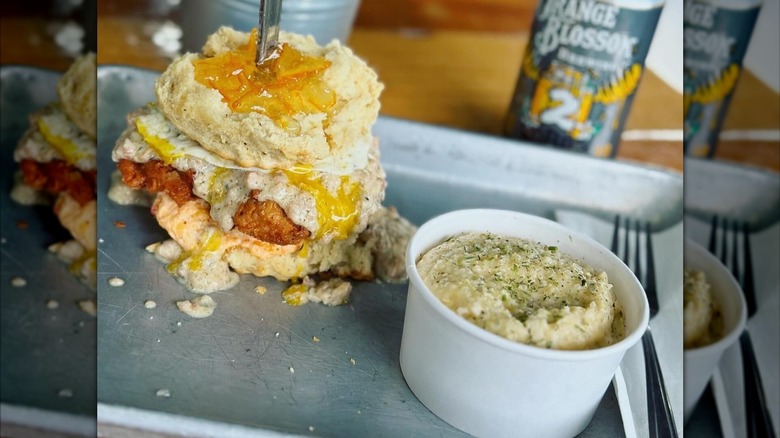 Fried chicken and egg on biscuit next to paper cup of grits on metal tray at Se7enBites
