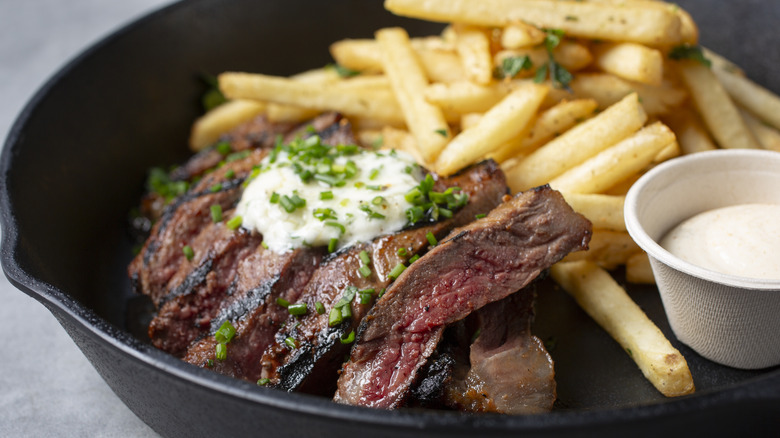 A skillet of steak frites with herb butter and a container of sauce