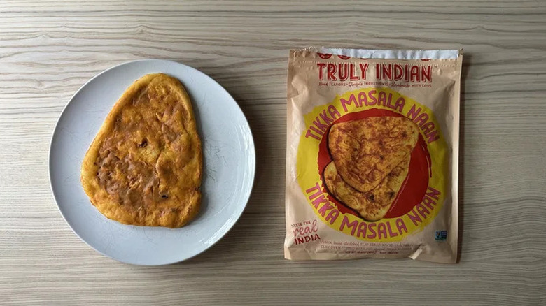 A Truly Indian Tikka Masala Naan on a white plate on a wooden surface, with its branded packaging sitting beside it.