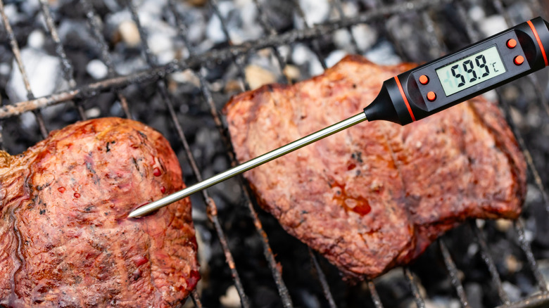 Someone using a meat thermometer on steaks cooking on a grill