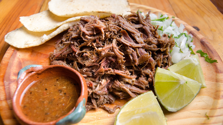 barbacoa with consome on a wood plate with tortillas and lime