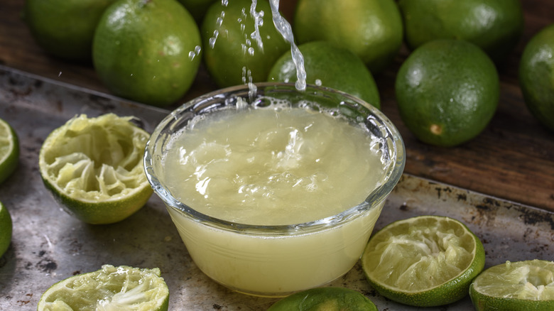 Lime juice being squeezed into a glass bowl, surrounded by several squeezed lime halves, and several whole limes in the background