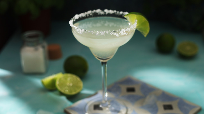 A salt-rimmed margarita with a wedge of lime, and several sliced limes sitting in the background