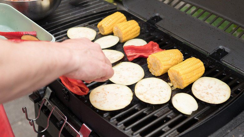 Hand placing eggplant slices on grill with other vegetables.