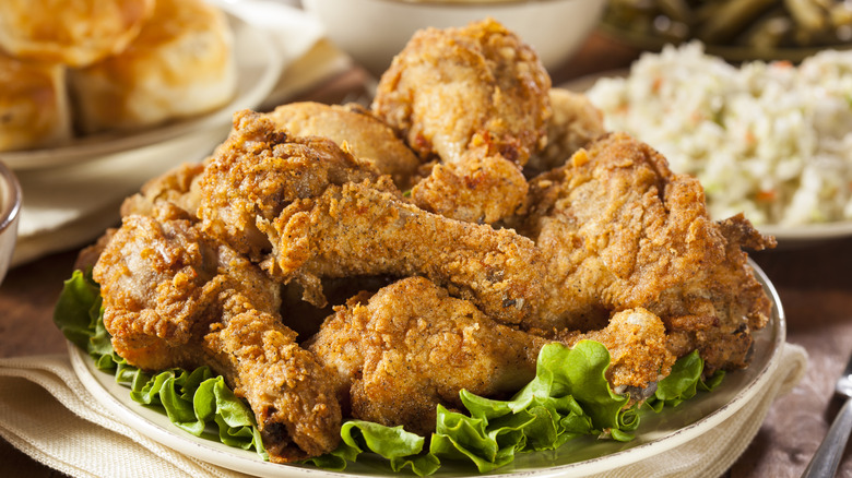 A white plate overloaded with crispy fried chicken, with coleslaw and biscuits in the background