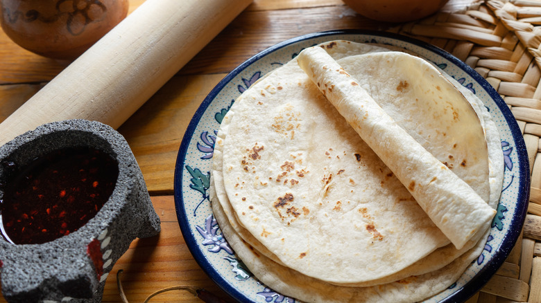 A blue and white plate holding a stack of fresh flour tortillas next to a container of salsa