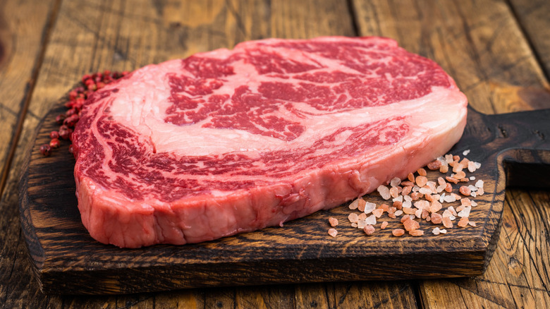a highly marbled raw ribeye steak on a cutting board with coarse pink salt and pink peppercorns