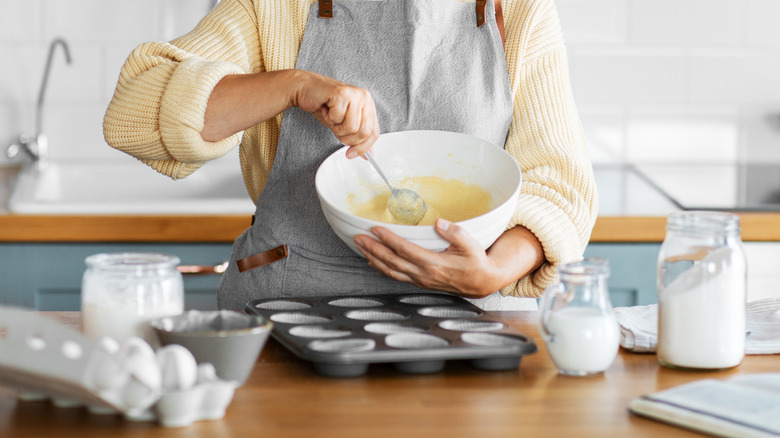 a person in a sweater and apron holding a mixing bowl and stirring batter with a spoon