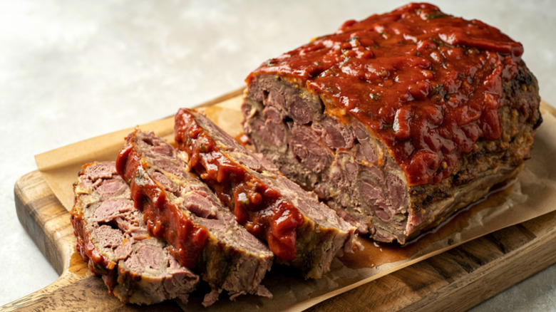 A partially sliced meatloaf covered in red sauce, sitting on a wooden cutting board