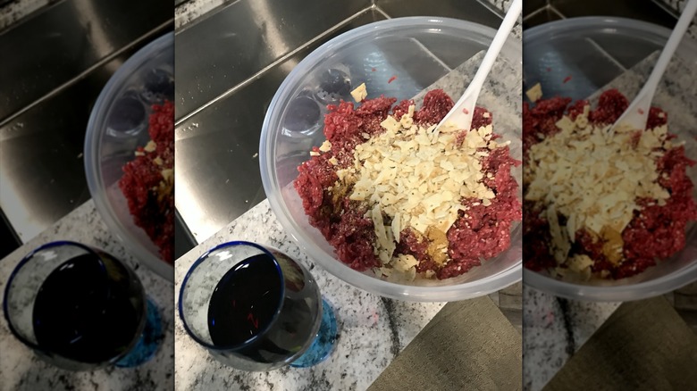 A glass of red wine next to a bowl of ground meat and crackers being turned into meatloaf