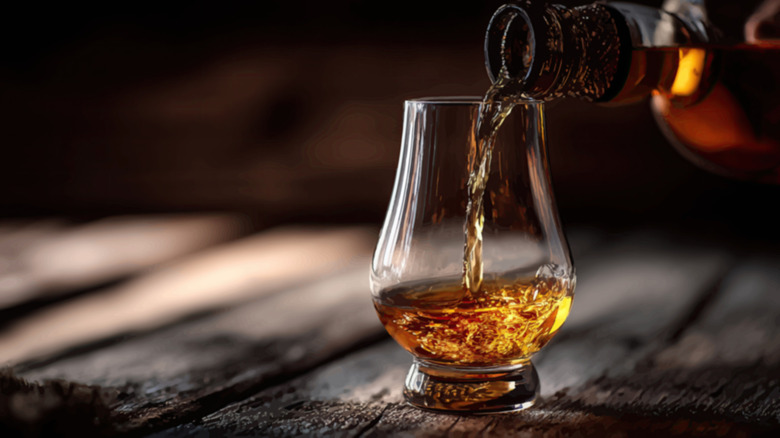 Bourbon pouring from a bottle into a tasting glass sitting on a wooden table