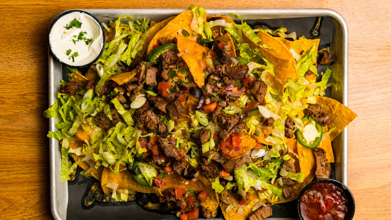 Brisket, lettuce, and tomato nachos on a metal sheet pan on top of a wooden table.