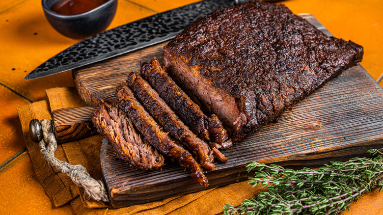 sliced brisket on a butting board with fresh thyme, a knife, and barbecue sauce