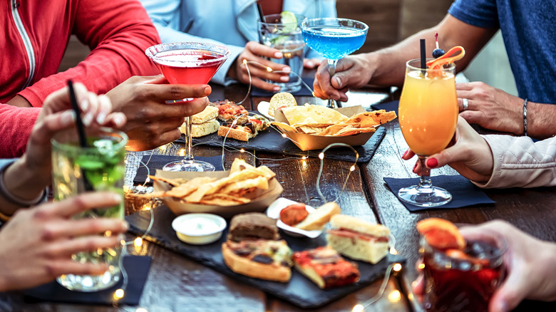 People seated around table filled with cocktails and small plates of food, including nachos and sandwiches