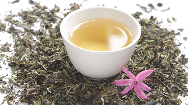A small cup of white tea surrounded by tea leaves and purple flower