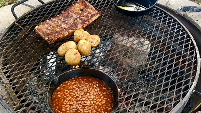 A cast iron skillet of beans, whole potatoes, and ribs cooking over a fire