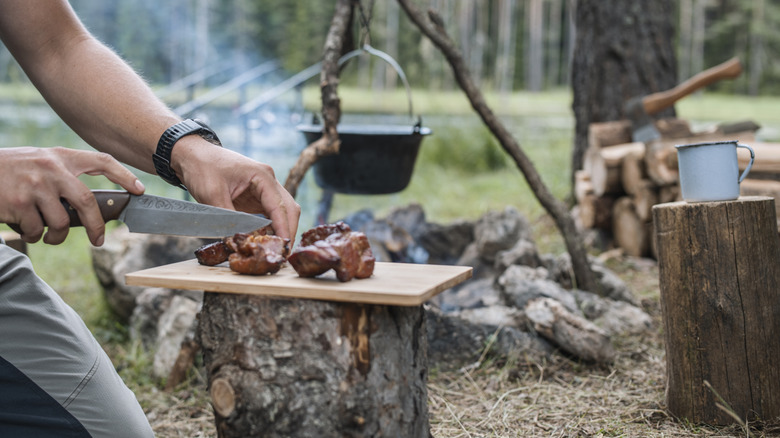 Someone cutting pieces of beef on a small board with a cast iron pot suspended over a fire in the background
