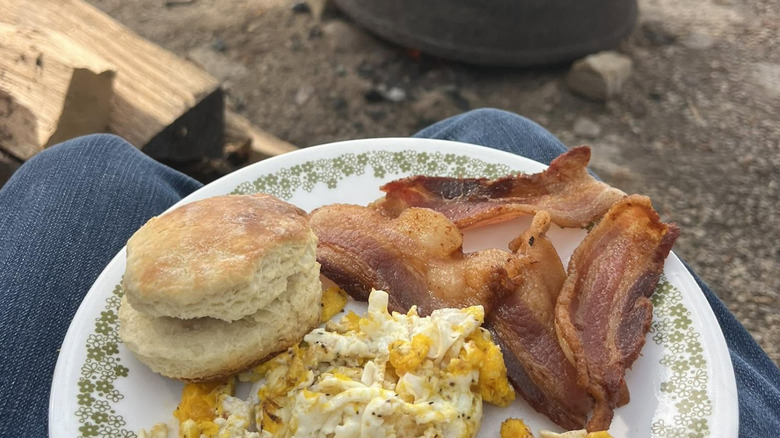 Bacon, eggs, and biscuits on plate beside campsite