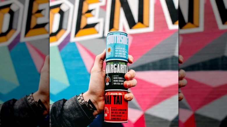 Hand holding three small cans of Post Meridiem canned cocktails