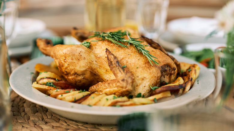 A whole roast chicken on a plate with some sliced potatoes and garnished with rosemary