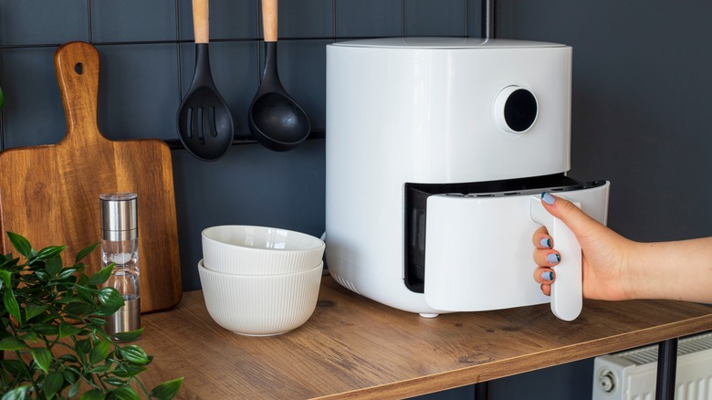 Woman holding the handle of a white air fryer, pulling out the basket