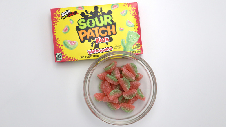 Watermelon Sour Patch Kids sit in a glass bowl on a white table with the box next to them