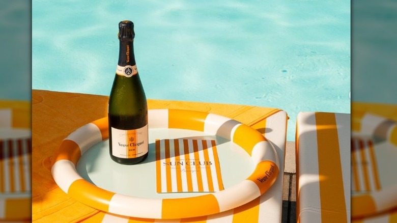 A bottle of Veuve Clicquot with its iconic bright logo sits on a pool float next to a pool