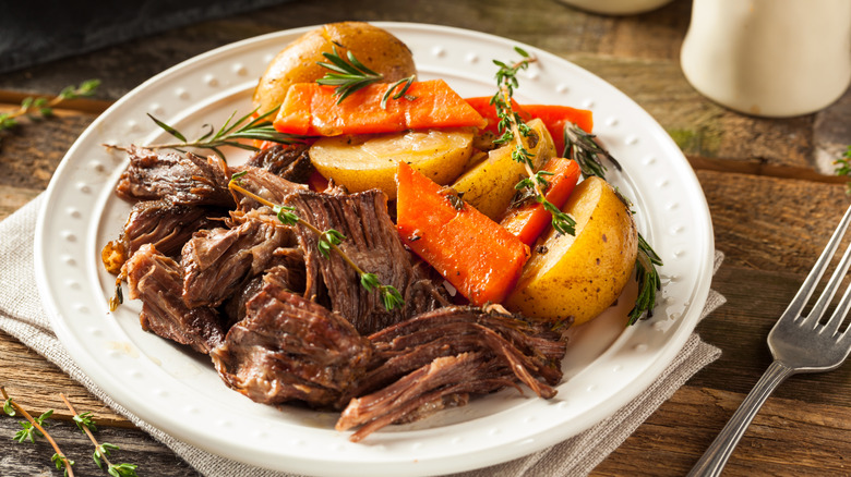 A plate of slow-cooker pot roast with veggies sitting next to a fork on a wooden table