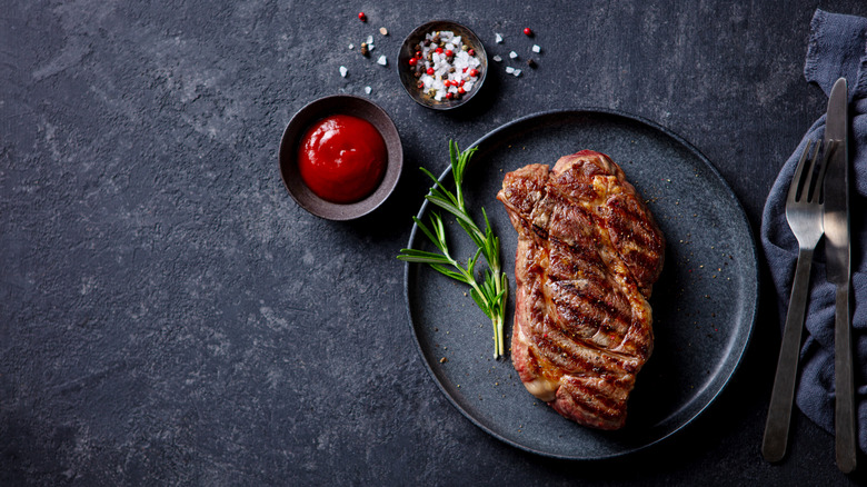 Steak cut on a plate with silverware, salt, and side of ketchup.