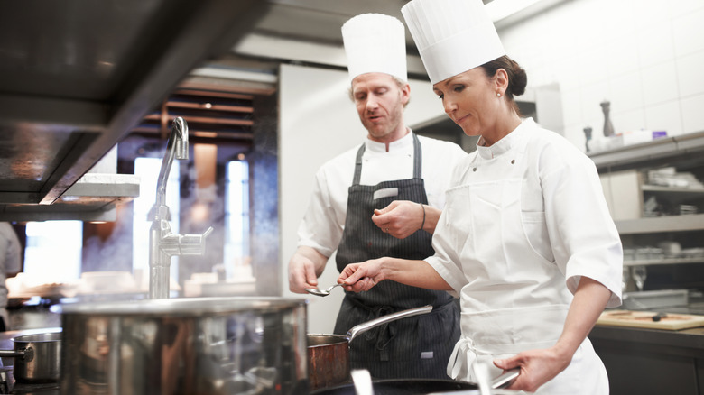 Two chefs watch over a boiling pot on a stove in a restaurant kitchen.