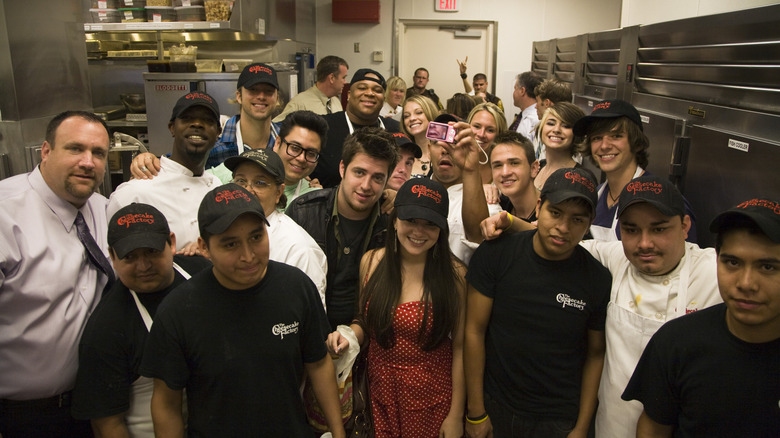 A collection of people in a kitchen are wearing branded ball caps with the Cheesecake Factory logo.