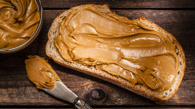 A piece of toast spread with peanut butter sitting on a wooden table next to a bowl of peanut butter and a spreader also covered in peanut butter